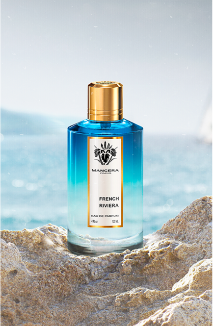 A blue bottle of Mancera Paris French Riviera 120ml Eau De Parfum with a gold cap is positioned among rocky terrain with a blurred ocean background.