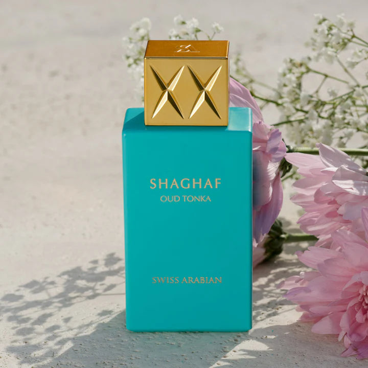 The turquoise bottle of Swiss Arabian Oud Tonka 75ml Eau De Parfum by Rio Perfumes, featuring a gold cap, stands elegantly beside pink flowers on a textured surface, capturing the essence of luxury. Buy 1 Get 1 Free offer available!.