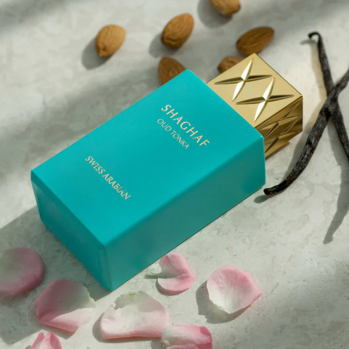 A turquoise "Swiss Arabian Oud Tonka 75ml Eau De Parfum" bottle with a gold cap, labeled Rio Perfumes, is elegantly arranged with almond nuts, vanilla pods, and pink rose petals on a textured surface. Buy 1 Get 1 Free offer available.