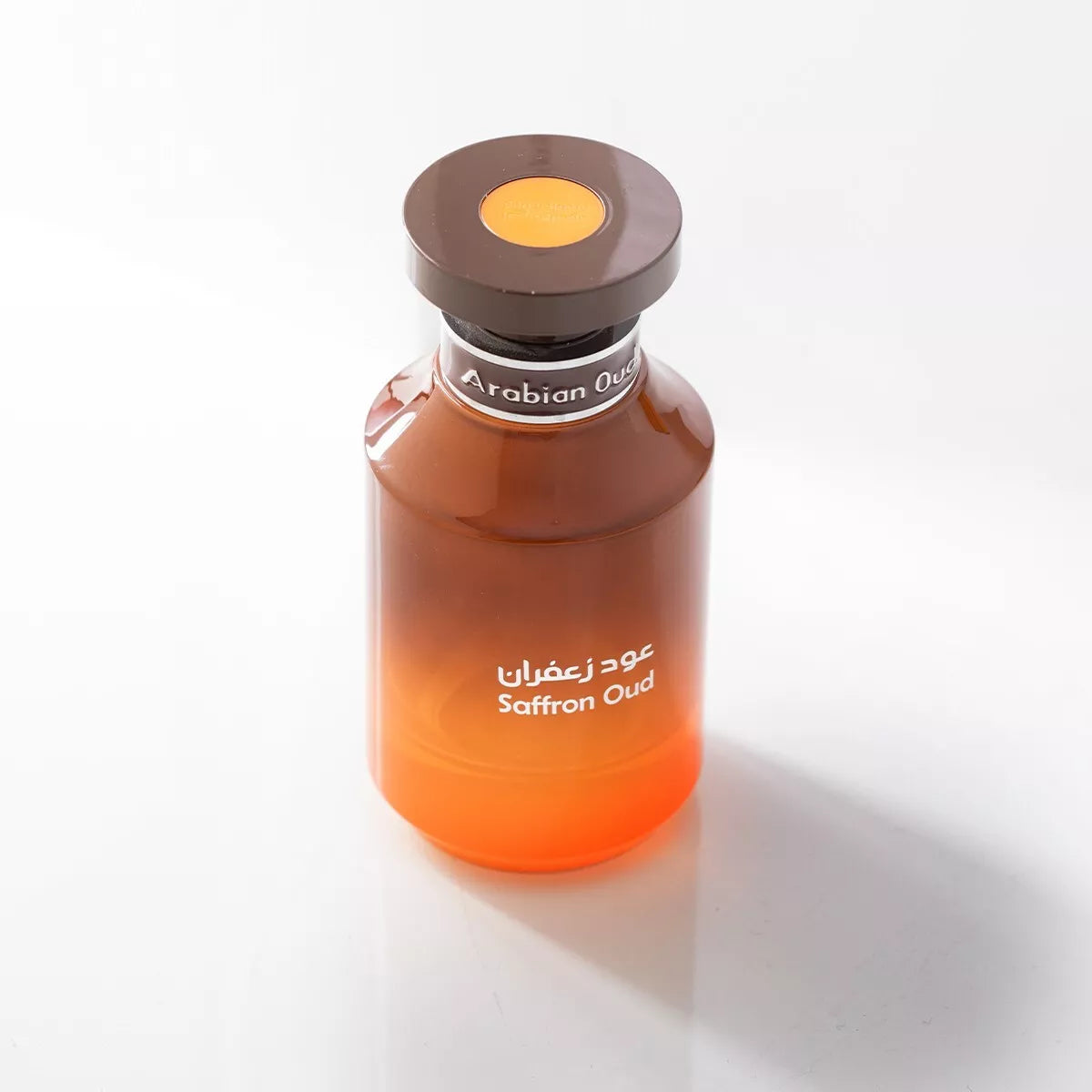 The Rio Perfumes Arabian Oud Saffron Oud 100ml Eau De Parfum, housed in a glass bottle with a gradient from dark to light amber and topped with a dark brown cap, presents an elegant Fragrance for Men against a white background.