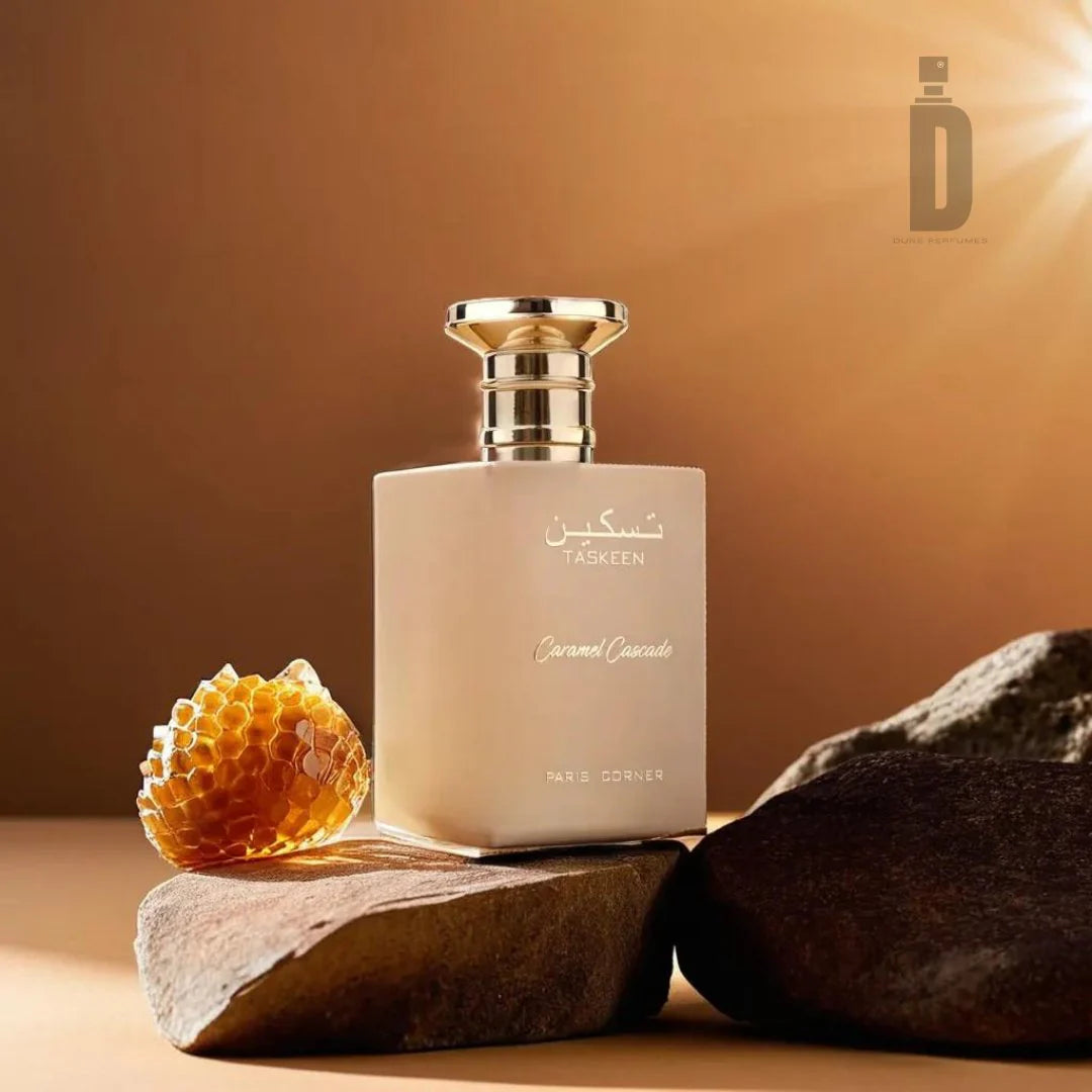 A beige perfume bottle labeled "Paris Corner Taskeen Caramel Cascade" is showcased on rocks beside a piece of honeycomb. Embellished with gold accents, the bottle is illuminated from above, enhancing its Floral Fruity Gourmand fragrance by Paris Corner.