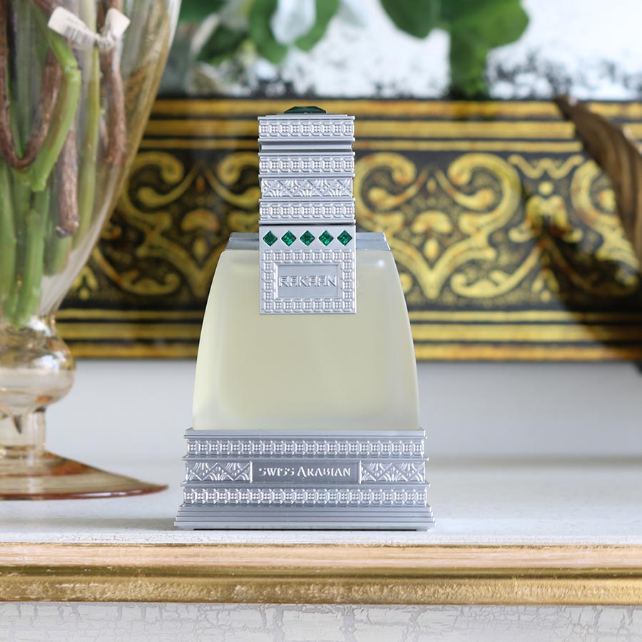 The Swiss Arabian Rakaan Eau De Parfum, featuring intricate silver detailing and emerald accents, exudes an intense woody fragrance. It's artfully placed on a white surface against a decorative background. Buy 1 Get 1 Free offer available.